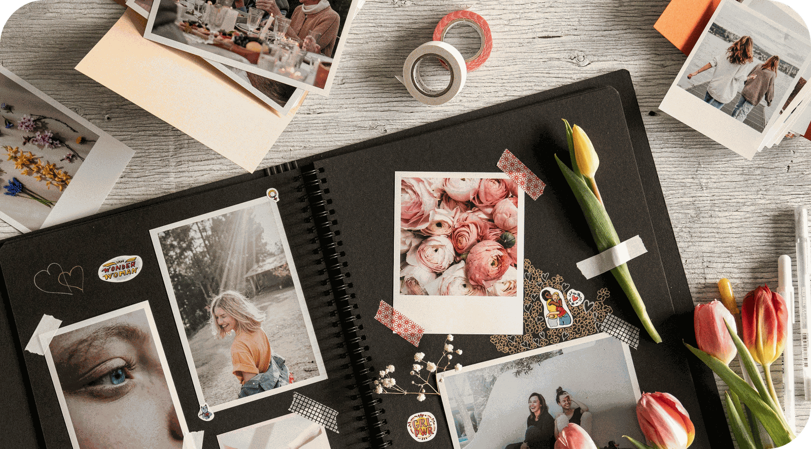 A flatlay of printed photographs, a photo album and flowers on a wooden desk
